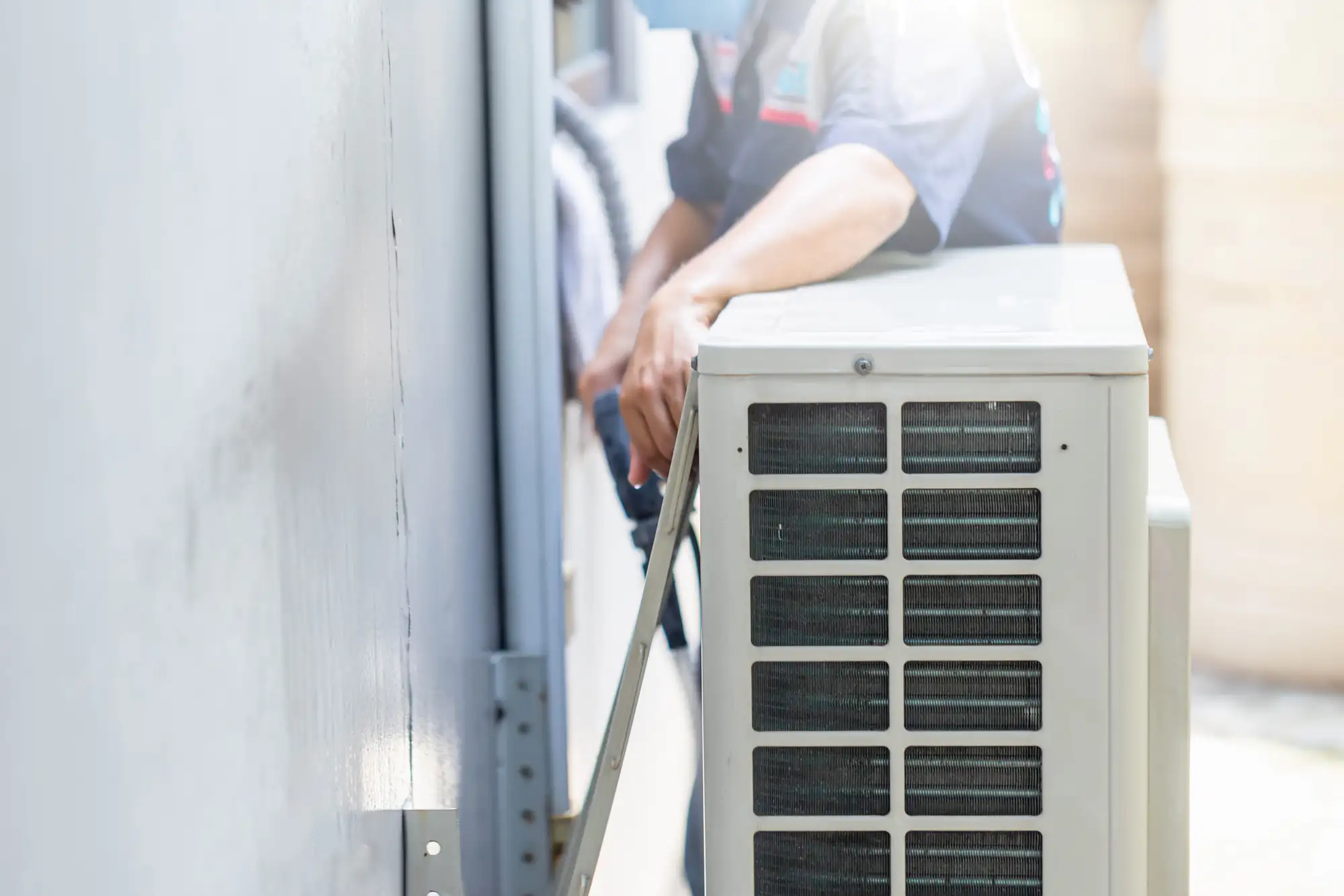 A technician in TX installs or repairs an outdoor air conditioning unit mounted against a wall, tools in hand and the unit's vented side visible—ideal for ac replacement Bexar County residents can rely on.
