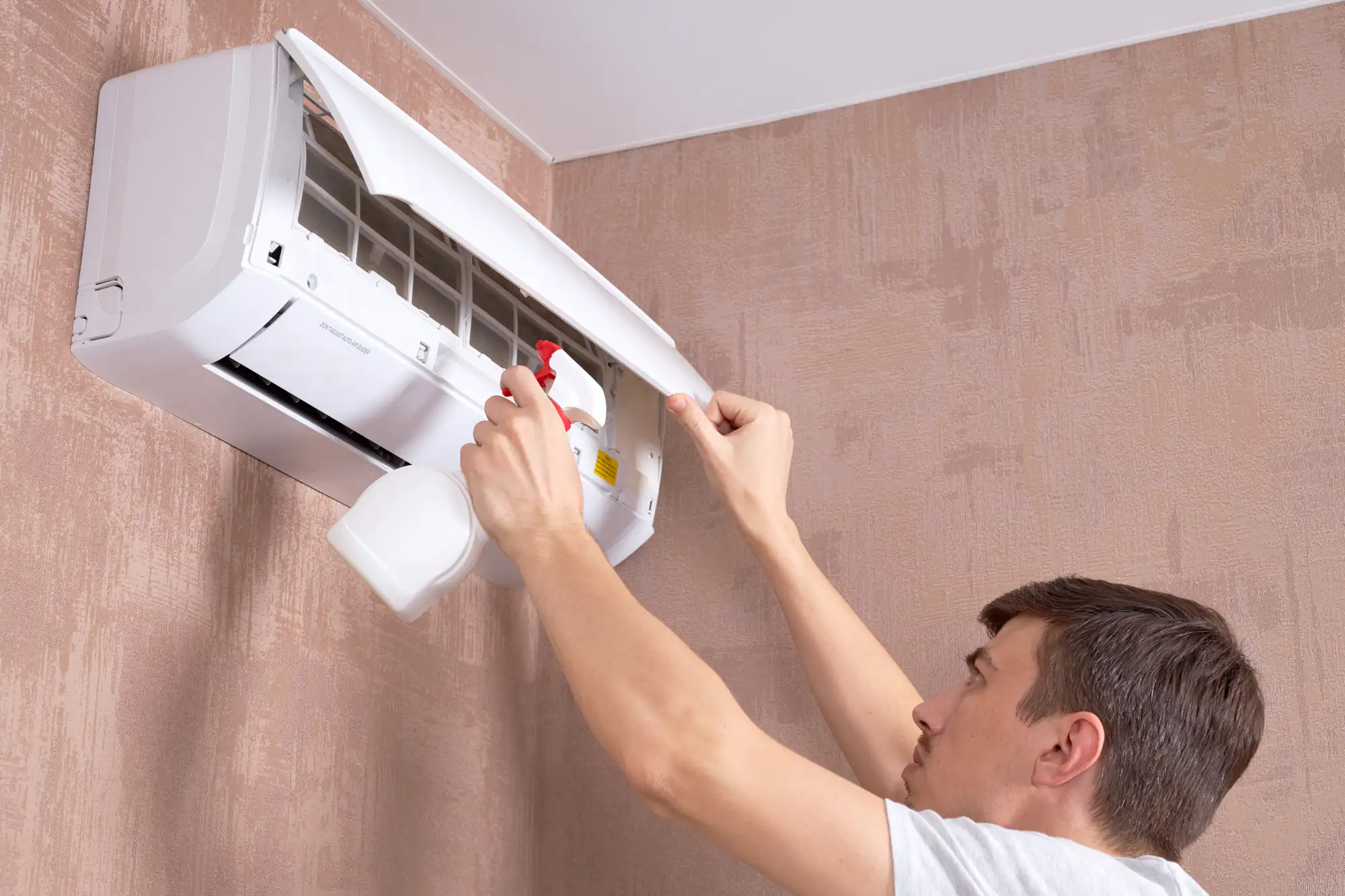 A man in TX inspects and cleans an open wall-mounted air conditioner with a spray bottle, standing close to a textured beige wall—essential maintenance before considering ac replacement Bexar County homeowners may need.