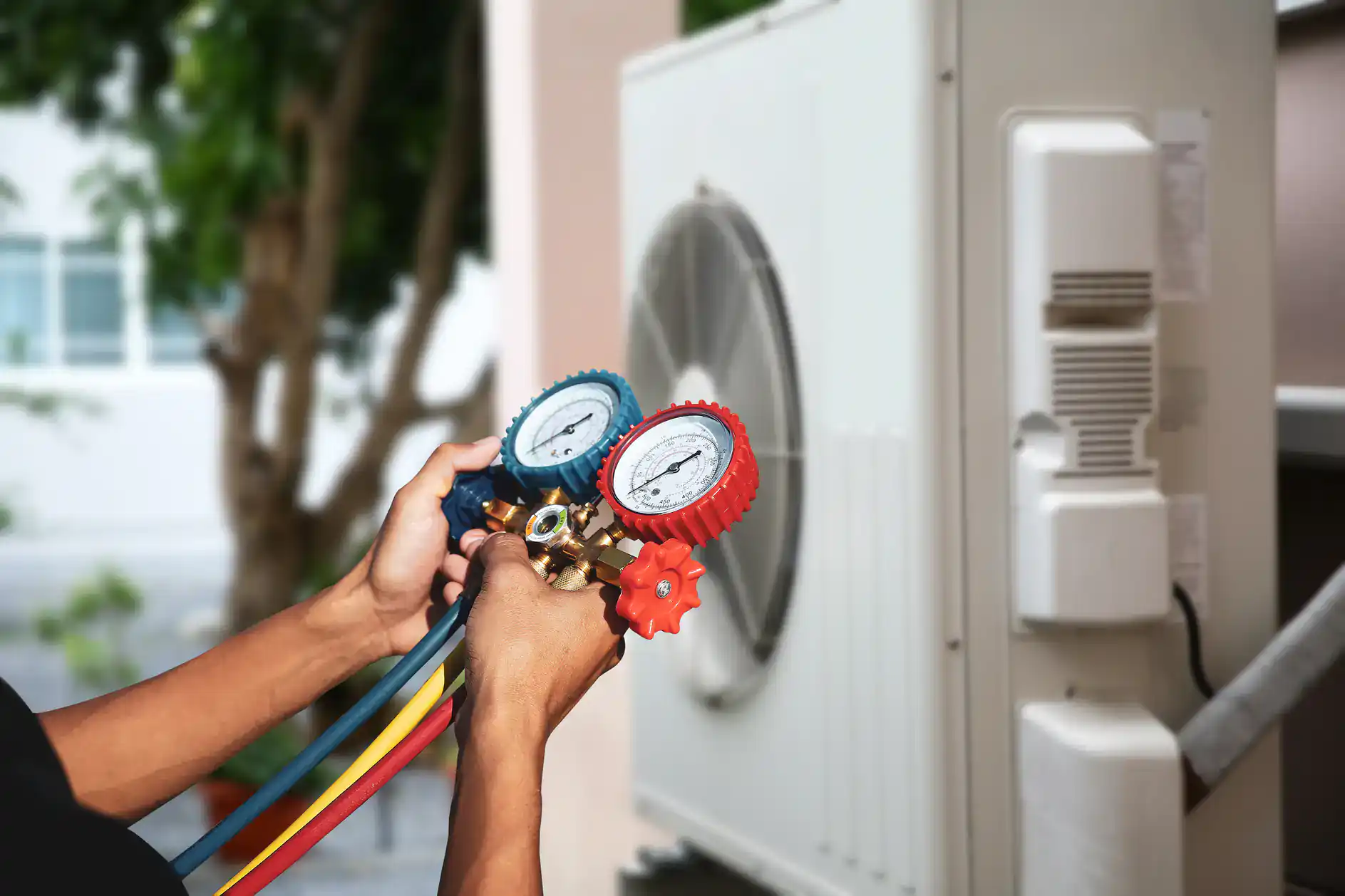 A person uses a manifold gauge set to check or service an outdoor air conditioning unit, possibly as part of an AC replacement in Bexar County, TX. The gauge has blue and red dials with colored hoses; a tree and part of a building are visible behind.