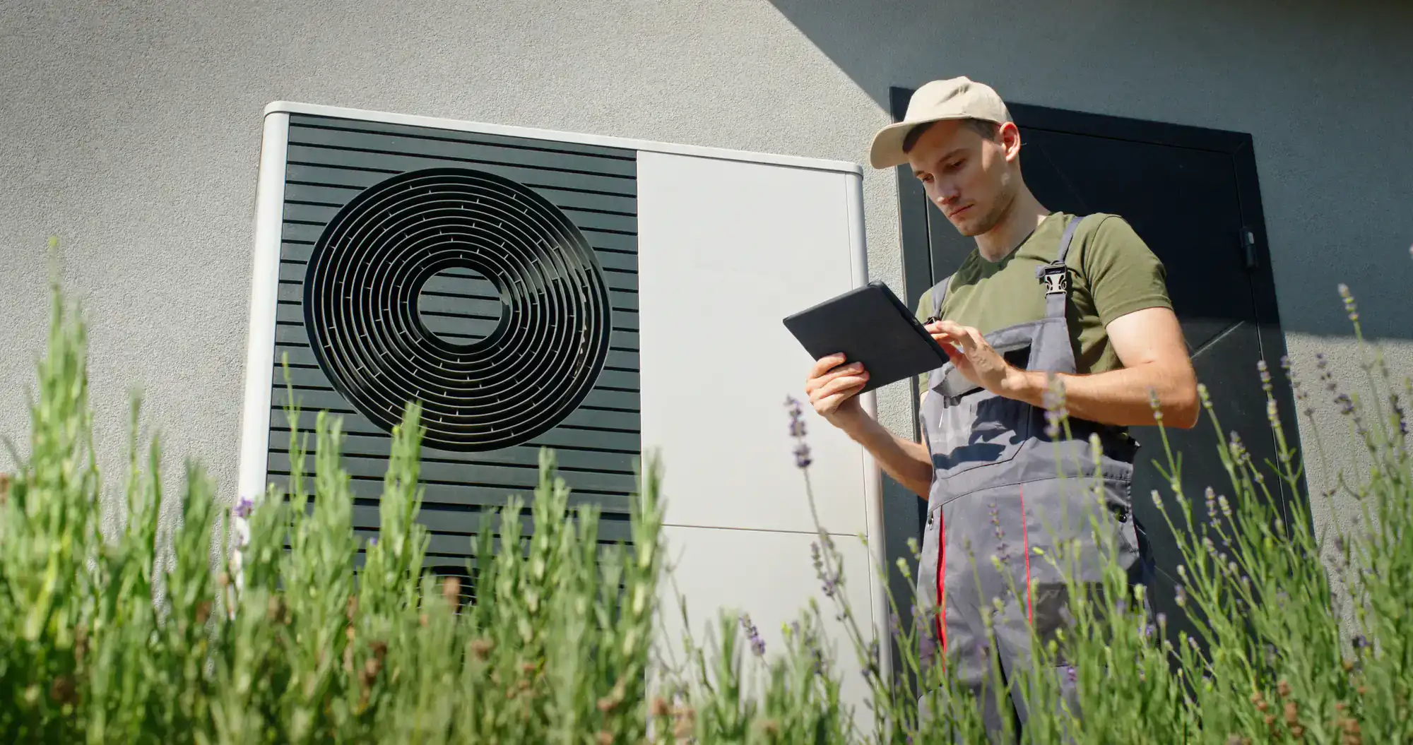 A technician in overalls and a cap stands outdoors in front of a heat pump unit in Bexar County, TX, using a digital tablet. Tall green plants are in the foreground, and a building wall is in the background—ideal for ac replacement projects.