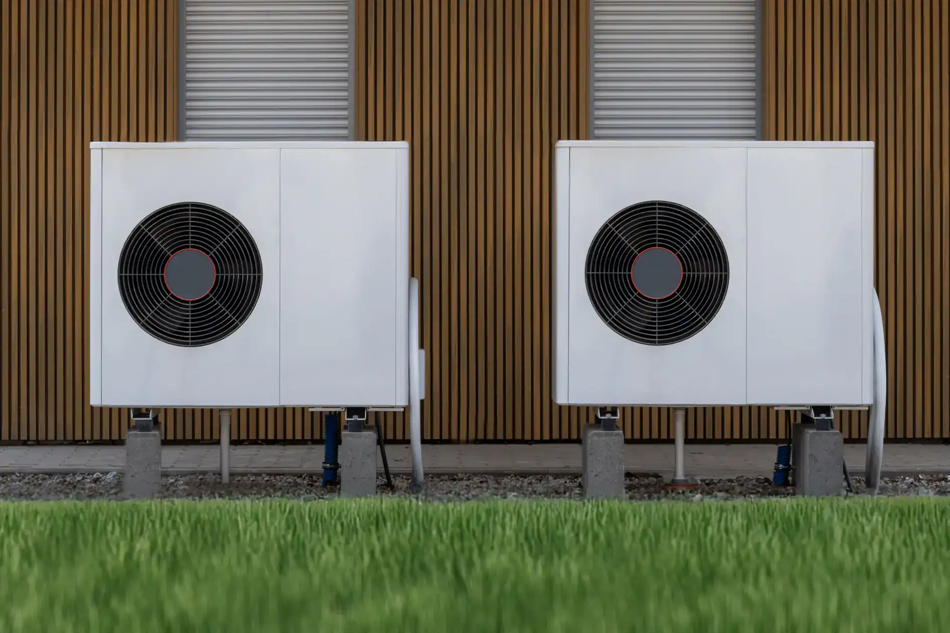 Two white outdoor heat pump units with large black fans stand side by side on concrete blocks in front of a modern building with vertical wooden slats and closed metal shutters in TX—ideal for ac replacement Bexar County residents can rely on. Green grass is visible in the foreground.