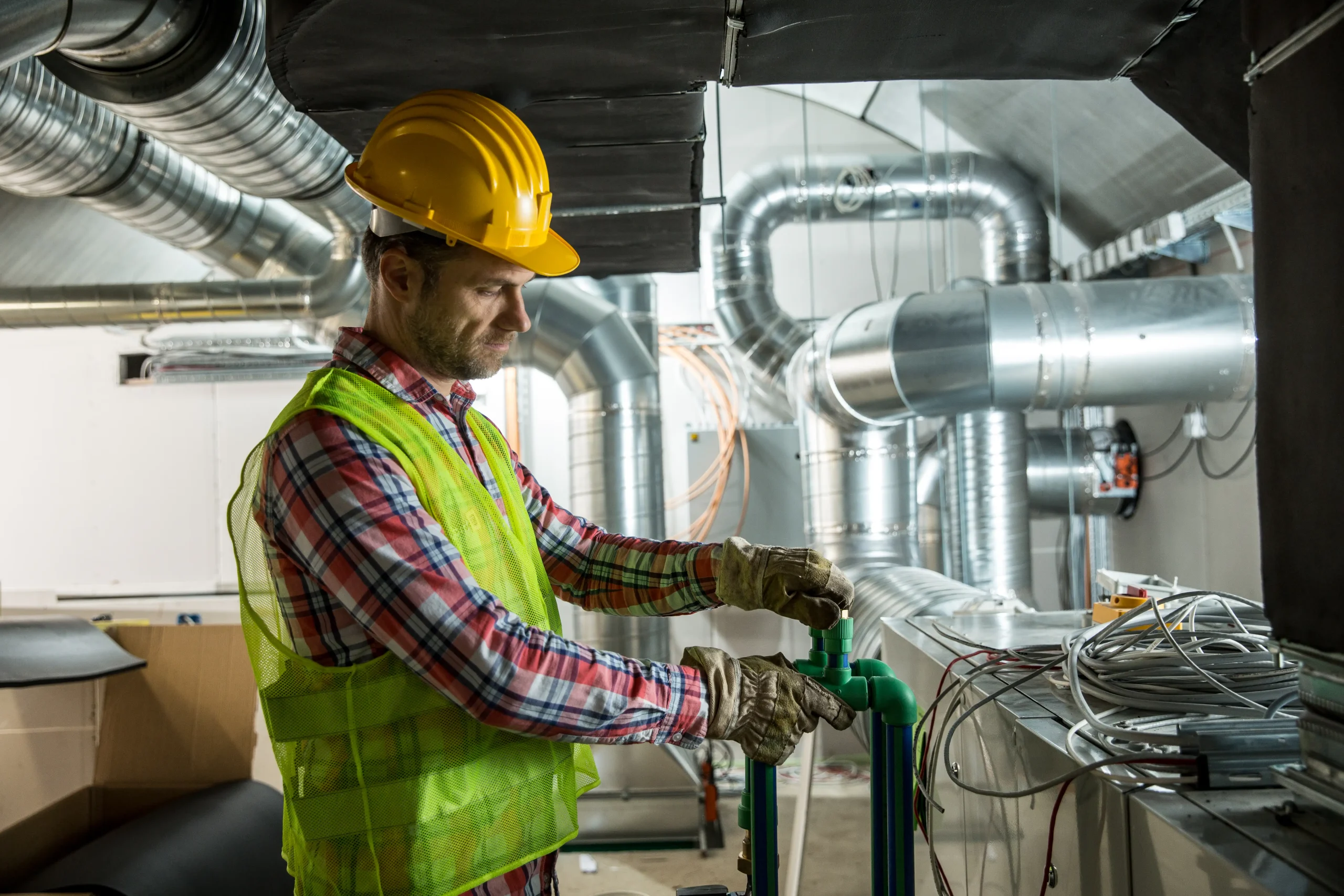 A construction worker in a yellow hard hat and safety vest adjusts green pipes in an industrial setting during an AC replacement in Bexar County, TX, with exposed metal ducts and various equipment in the background.