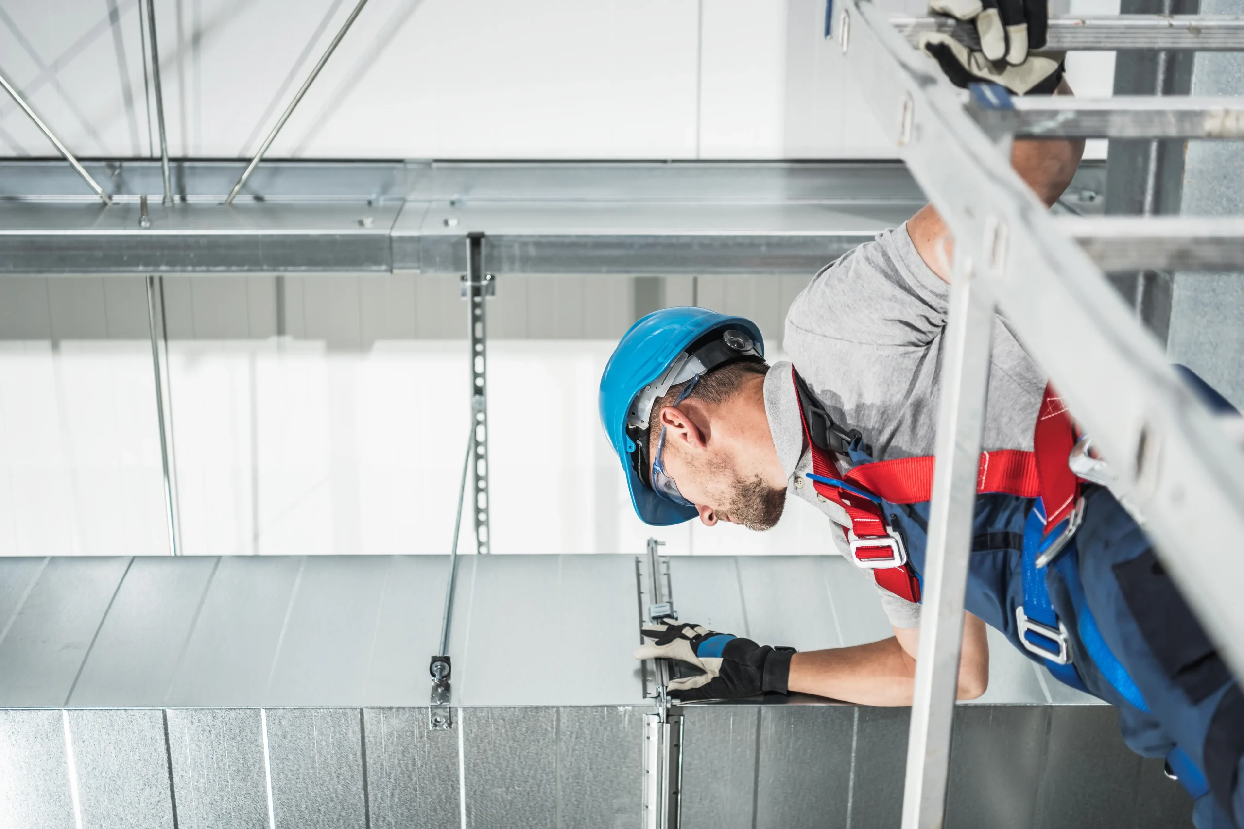 A construction worker wearing a blue hard hat, gloves, and a safety harness installs or inspects metal ductwork on a ceiling—possibly for an AC replacement Bexar County, TX project—while standing on a ladder inside an industrial or commercial building.