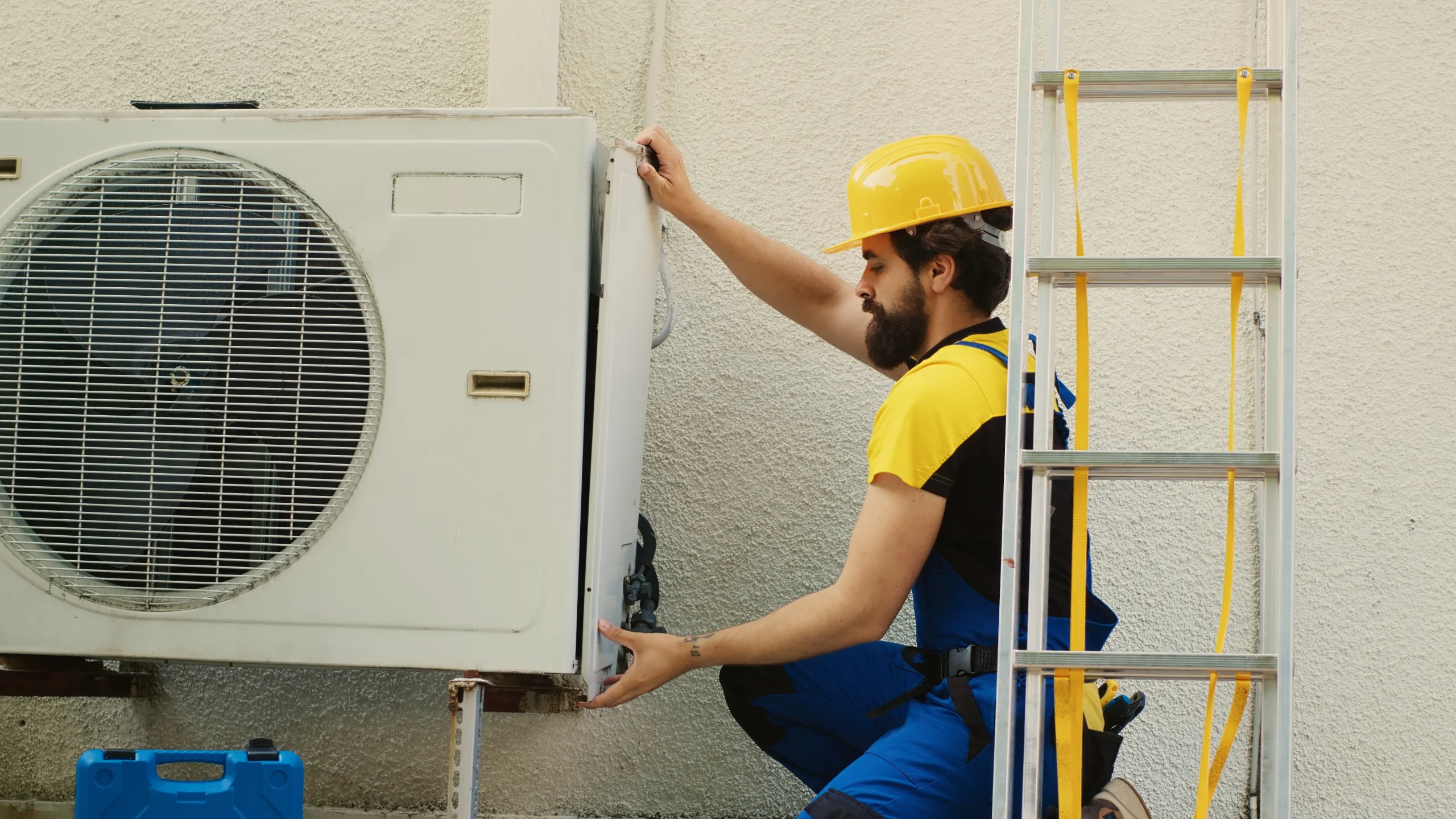 A worker in a yellow hard hat and blue overalls is kneeling beside a yellow ladder, installing or repairing an outdoor air conditioning unit in Bexar County, TX, possibly as part of an AC replacement job.