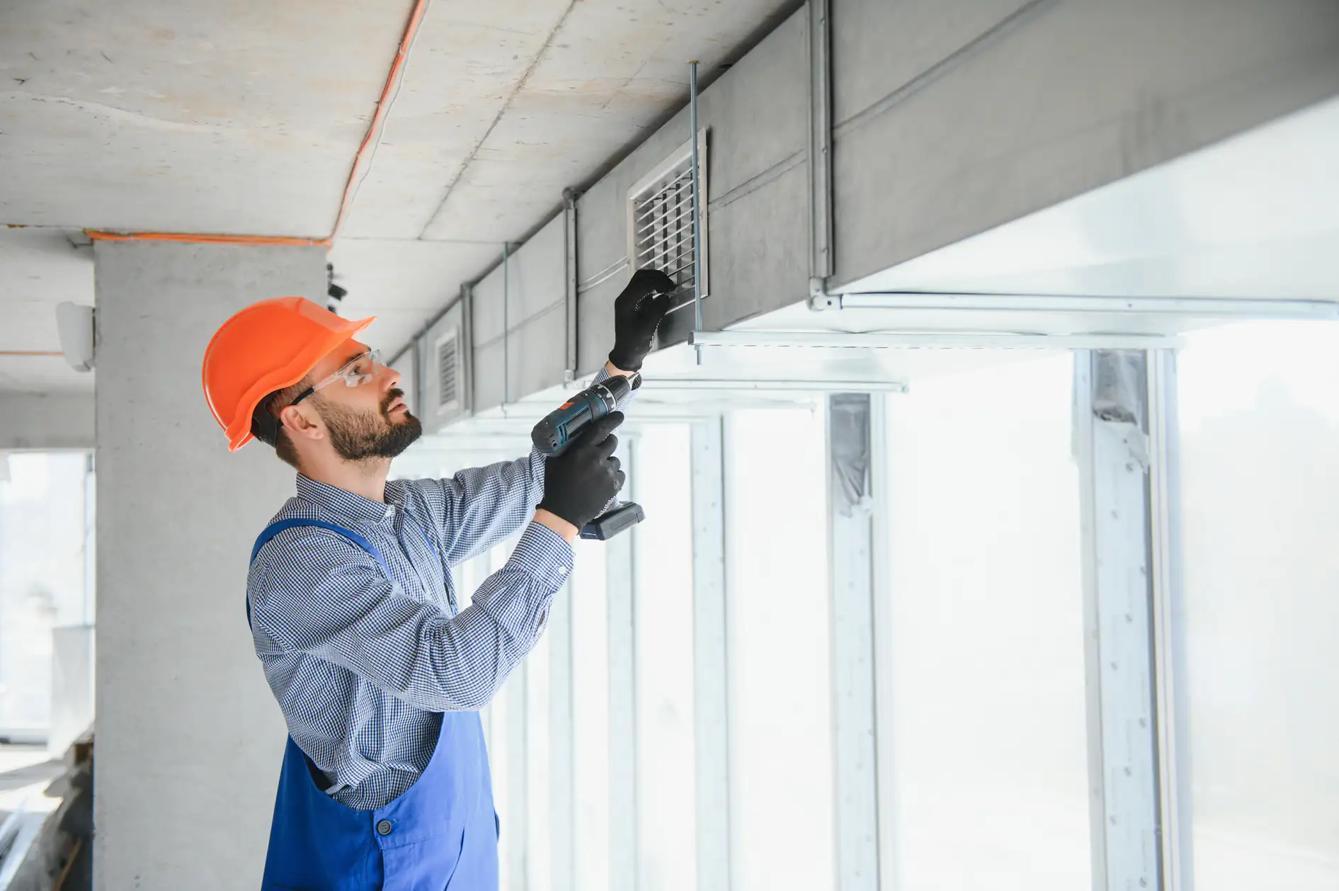 A construction worker in a hard hat and gloves uses a power drill to install or repair a ventilation grate on a ceiling duct, part of an AC replacement project in Bexar County, TX, inside a bright, modern building.