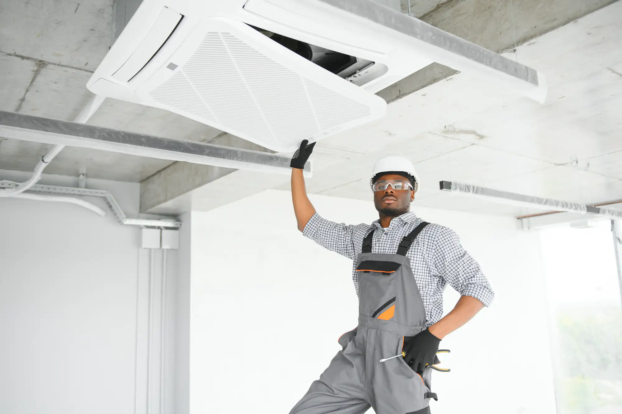 A technician in overalls, safety glasses, gloves, and a hard hat inspects or repairs a ceiling-mounted air conditioning unit in a modern, unfinished room—perfect for TX summers or ac replacement in Bexar County.