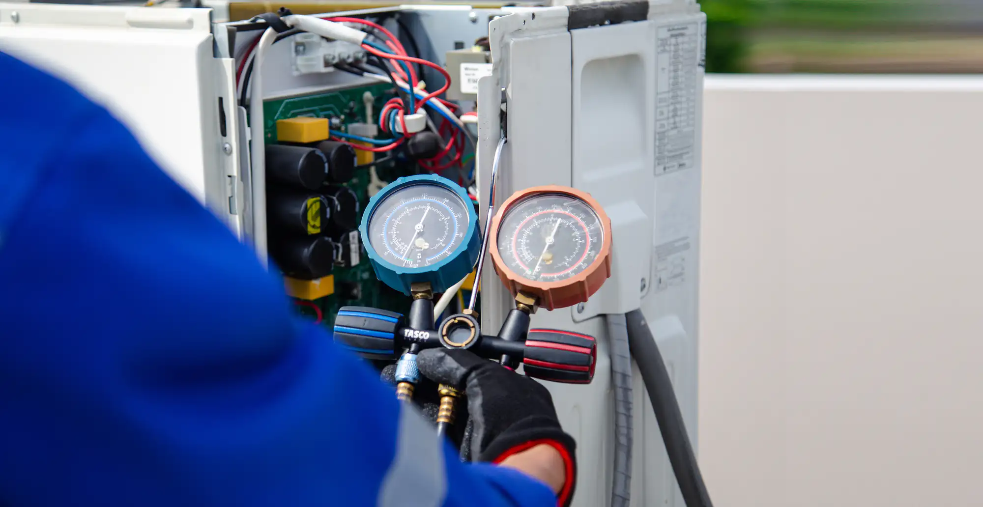 A technician in a blue uniform uses a manifold gauge to check the pressure on an open air conditioning unit with exposed wires and circuit board, ensuring the system is ready for ac replacement in Bexar County, TX.