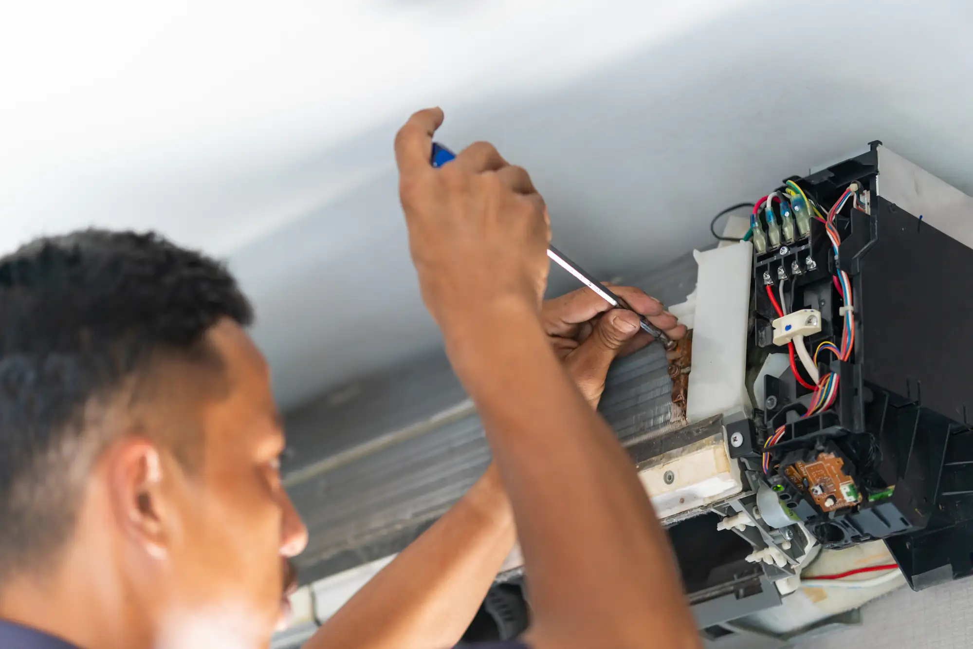 A person in TX uses a screwdriver to repair or maintain the internal components of a wall-mounted air conditioning unit, with exposed wires and parts visible—common during ac replacement Bexar County residents may need.