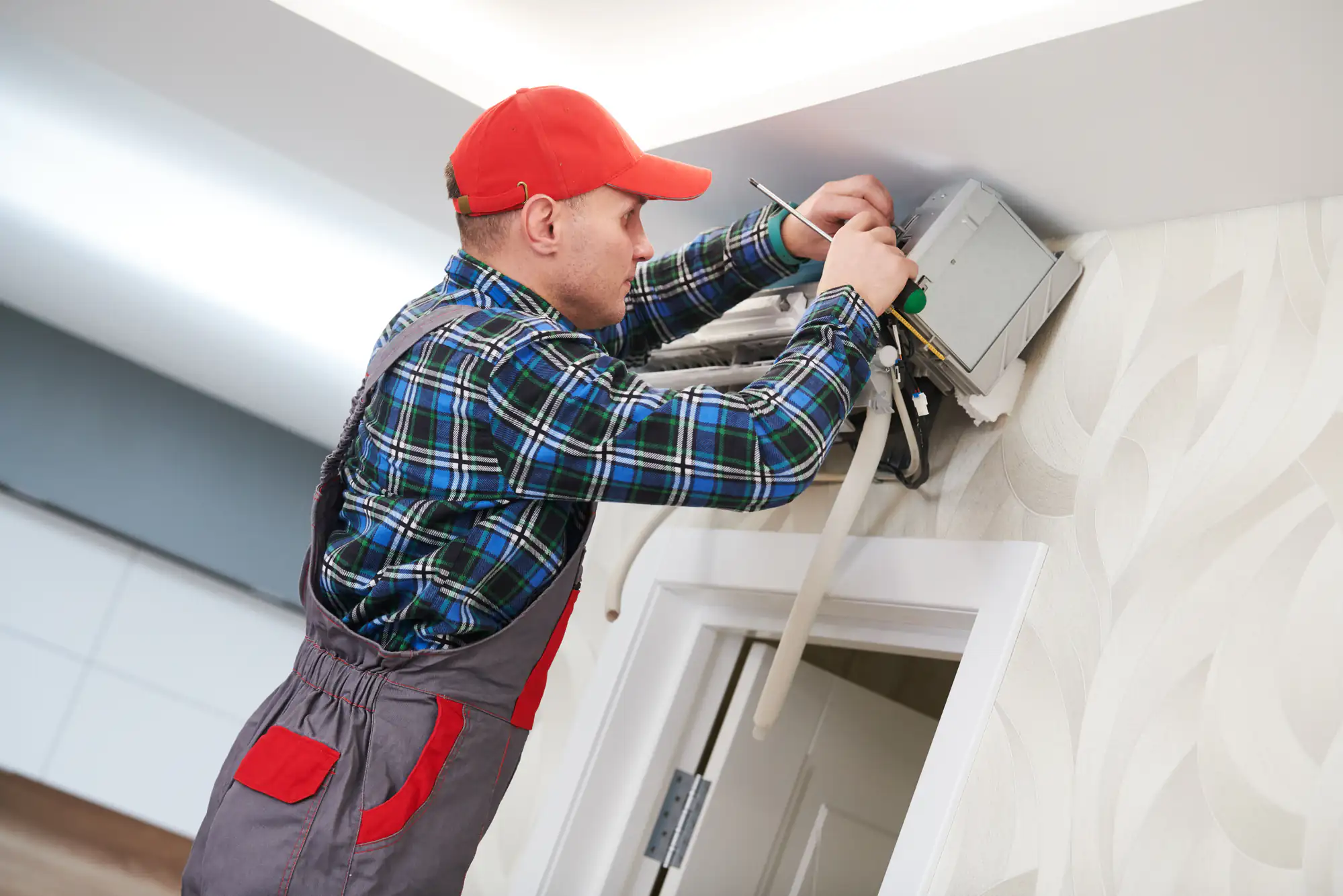 A technician wearing a red cap, plaid shirt, and overalls repairs an air conditioning unit mounted high on a wall inside a TX home, using tools and a screwdriver—ideal for AC replacement Bexar County residents may need.