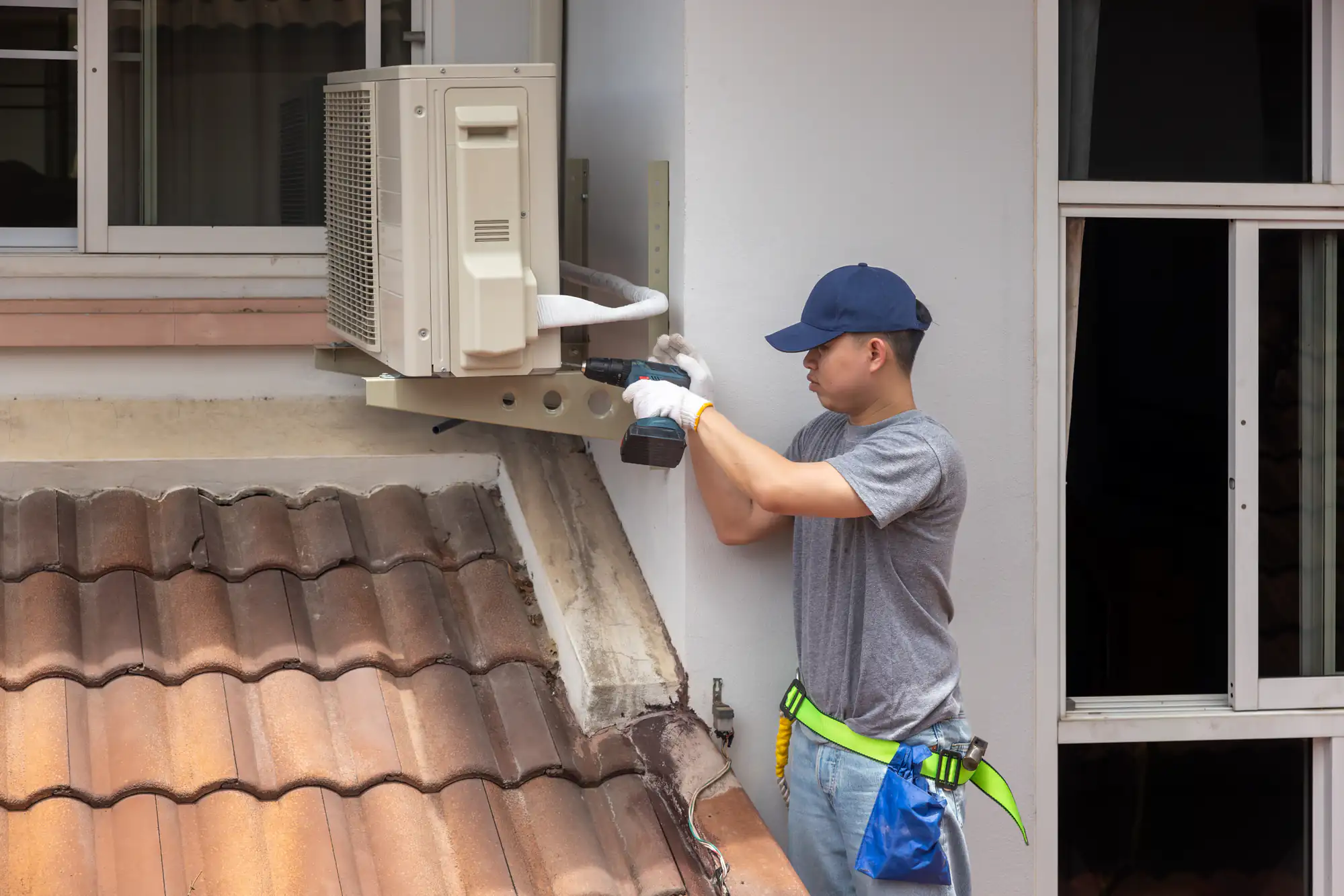A technician in a gray shirt and blue cap uses a power drill to install or repair an outdoor air conditioning unit above a tiled roof, showcasing expert ac replacement in Bexar County, TX. Tools hang from his belt, and a window is visible nearby.