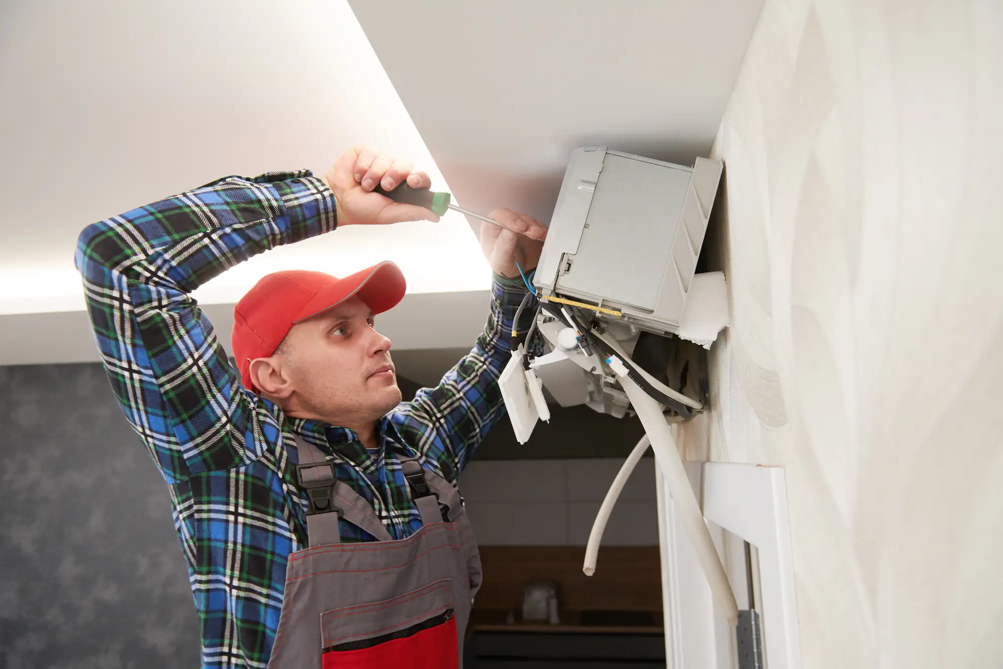 A technician in a red cap and plaid shirt uses a screwdriver to fix an air conditioning unit mounted on a wall, showcasing his expertise in AC replacement in Bexar County, TX. Cables and tools are visible as he works on the device.