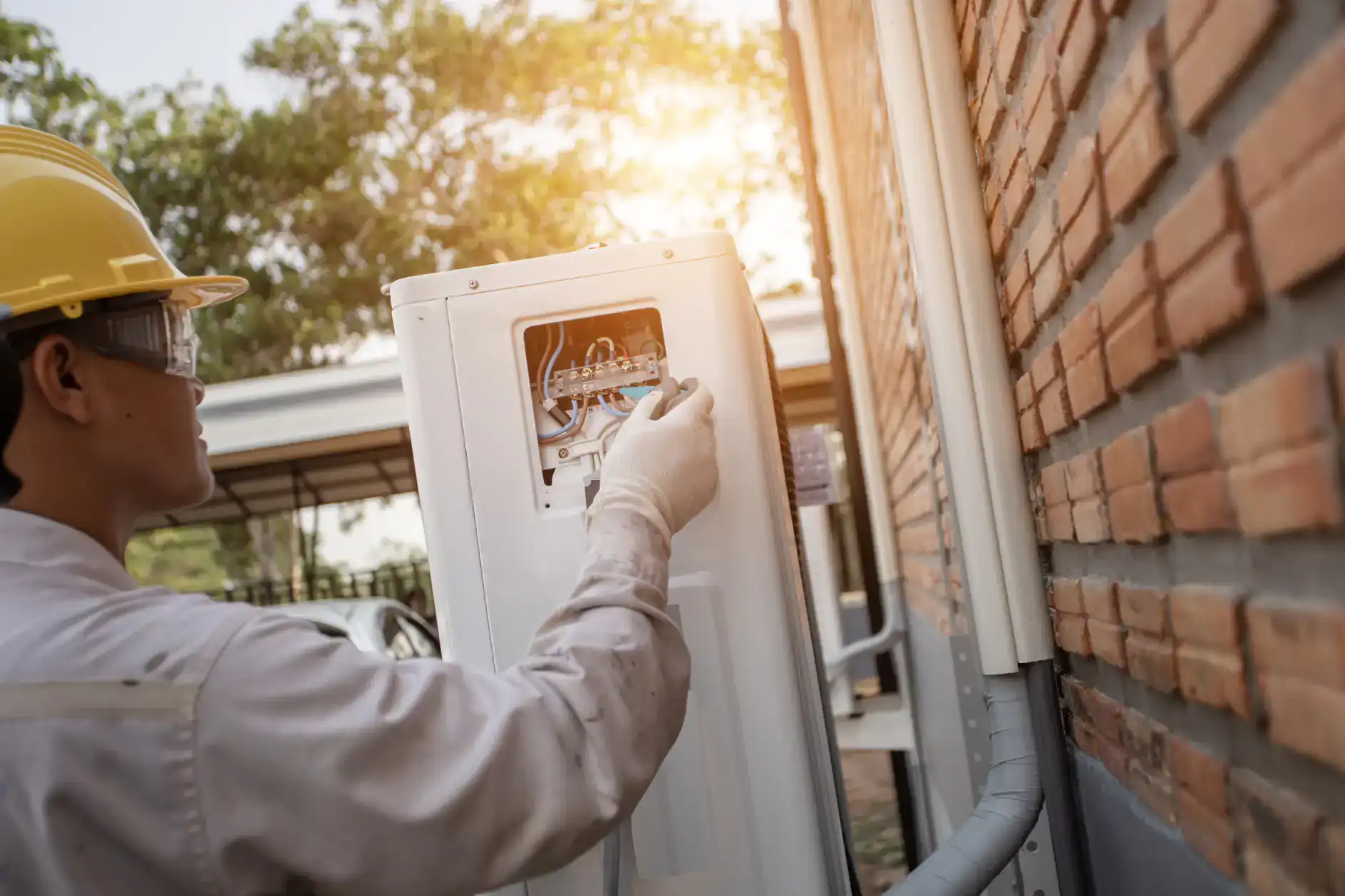 A worker wearing a yellow hard hat and white gloves repairs or installs an electrical unit on a building’s brick wall in Bexar County, TX, with sunlight shining in the background—possibly part of an ac replacement project.