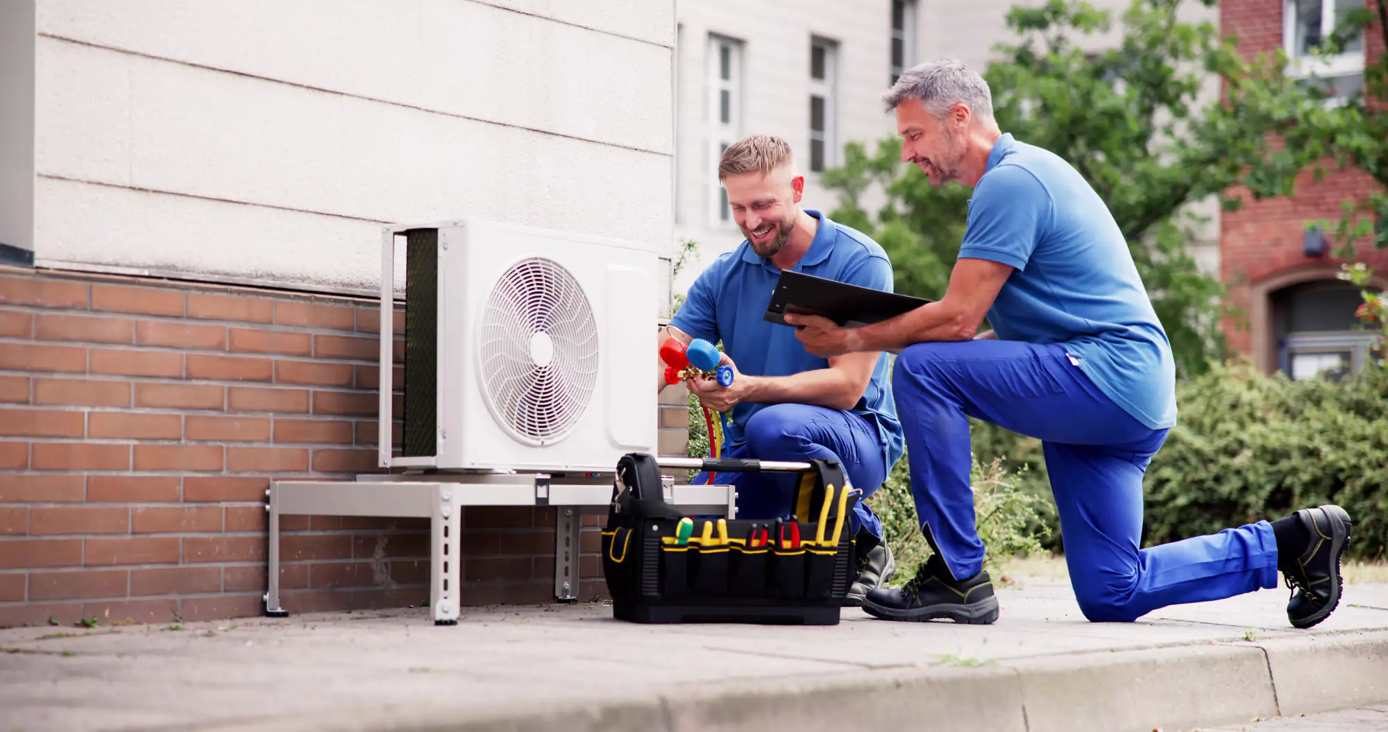 Two male HVAC technicians in blue uniforms work on an outdoor air conditioning unit beside a building in Bexar County, TX; one holds a clipboard while the other uses tools from a black toolbox for an AC replacement.
